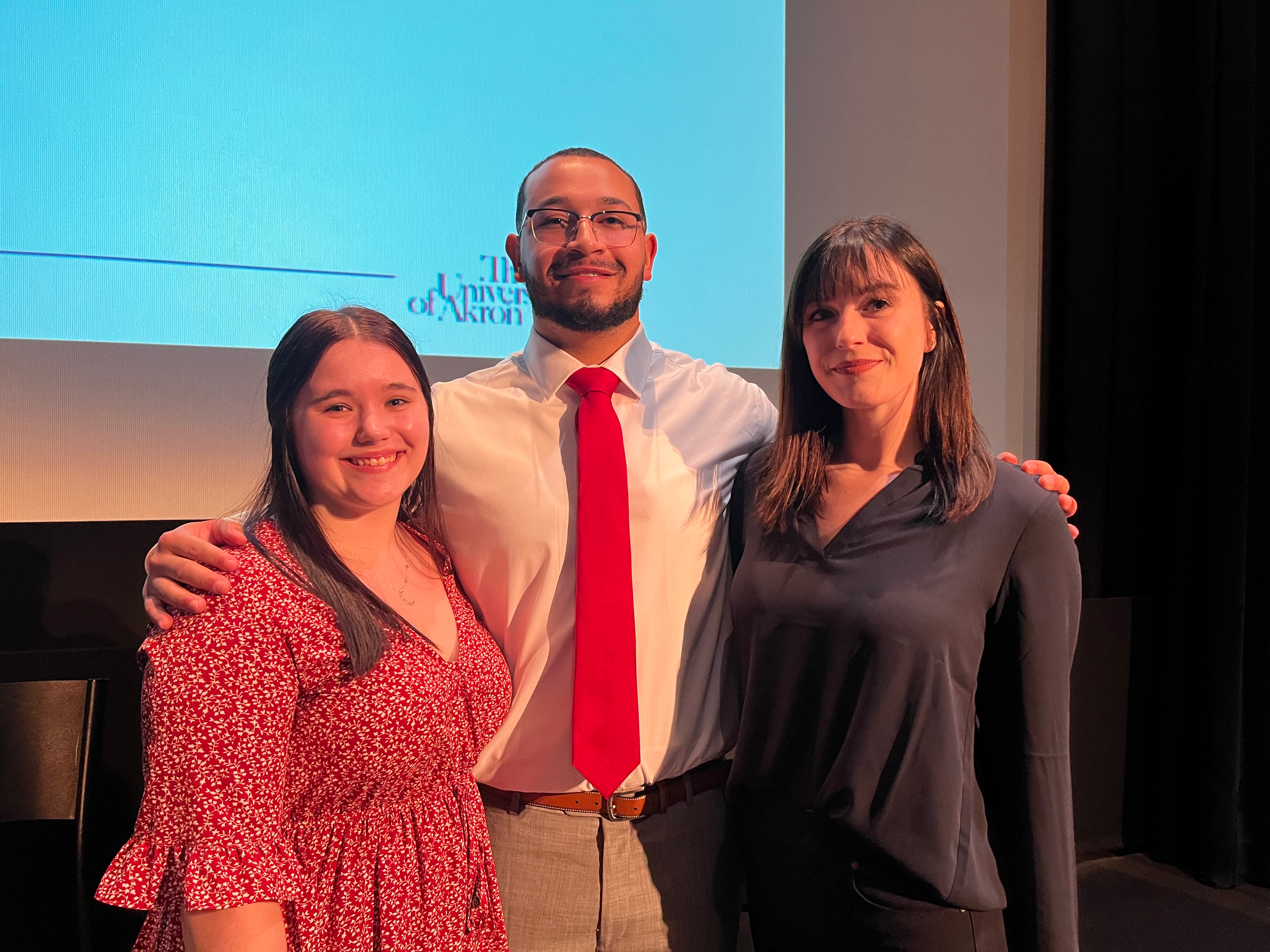 Three Akron students pose in front of an event presentation for the Ohio Innocence Project